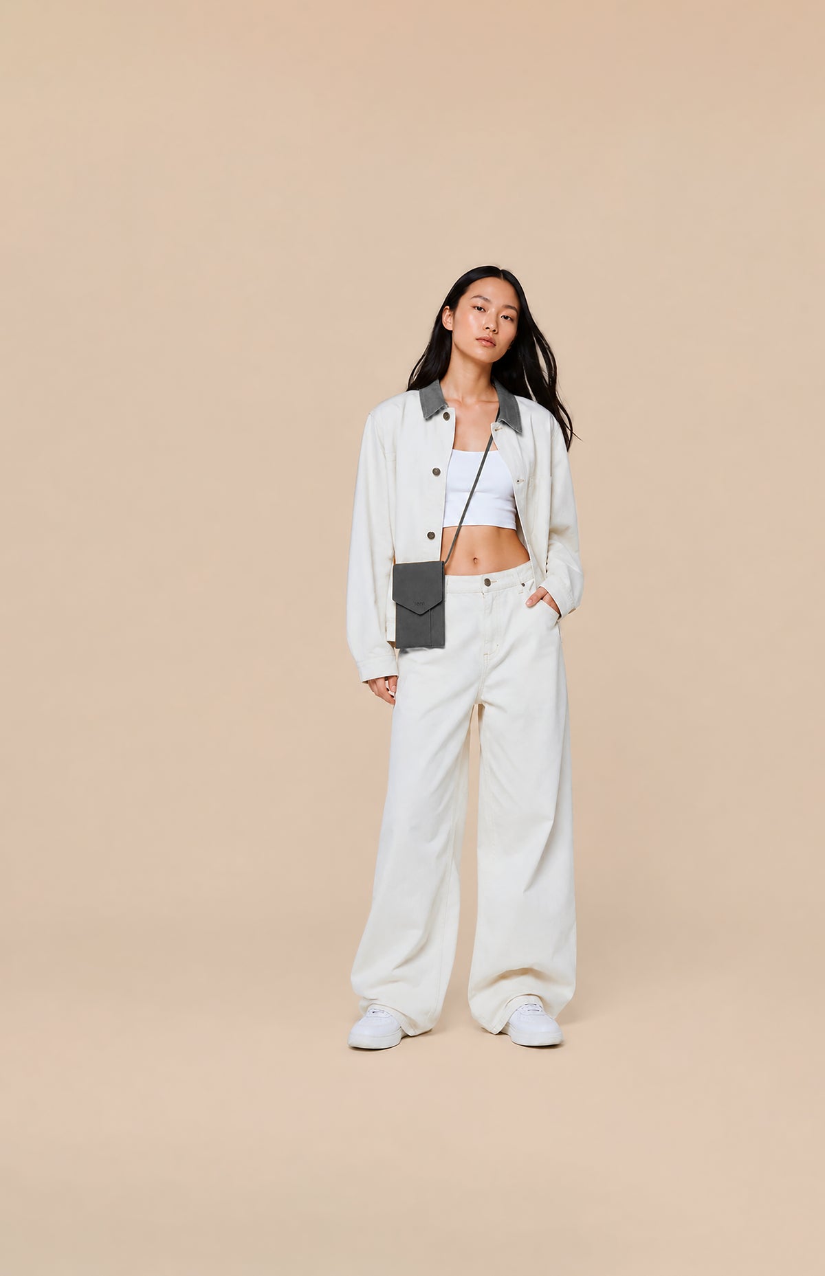 A model stands against a solid beige studio background wearing a white button-up shirt with dark gray collar and pocket details, matching wide-leg white trousers, a white crop top, and white sneakers. She is also wearing a small gray crossbody phone bag.